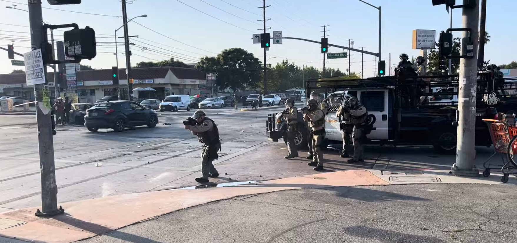 Sheriffs deputies in front of a sheriff's pick-up parked on the curb point their weapons at protesters off screen.