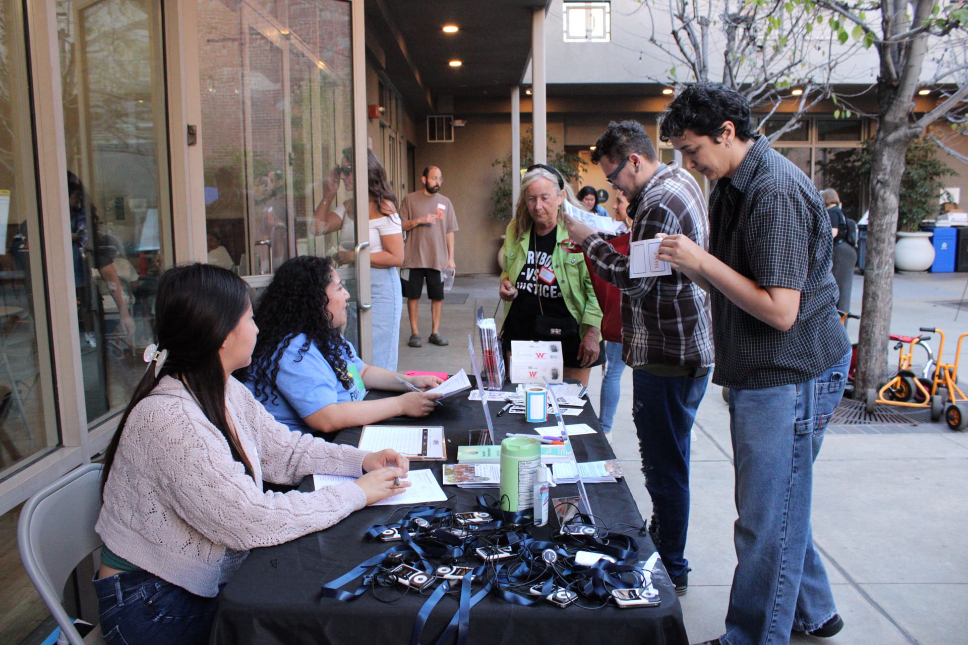 Five people, two sitting behind a table with various lanyards and flyers on it, interacting in a courtyard setting.