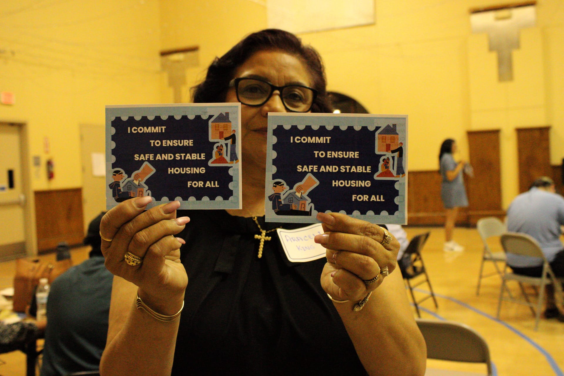 A woman with black-rimmed glasses holding up two signs that say "I commit to ensure safe and stable housing for all."